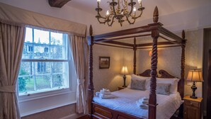 Una cama con dosel, una araña, una ventana con cortinas y una mesita de noche con lámpara.