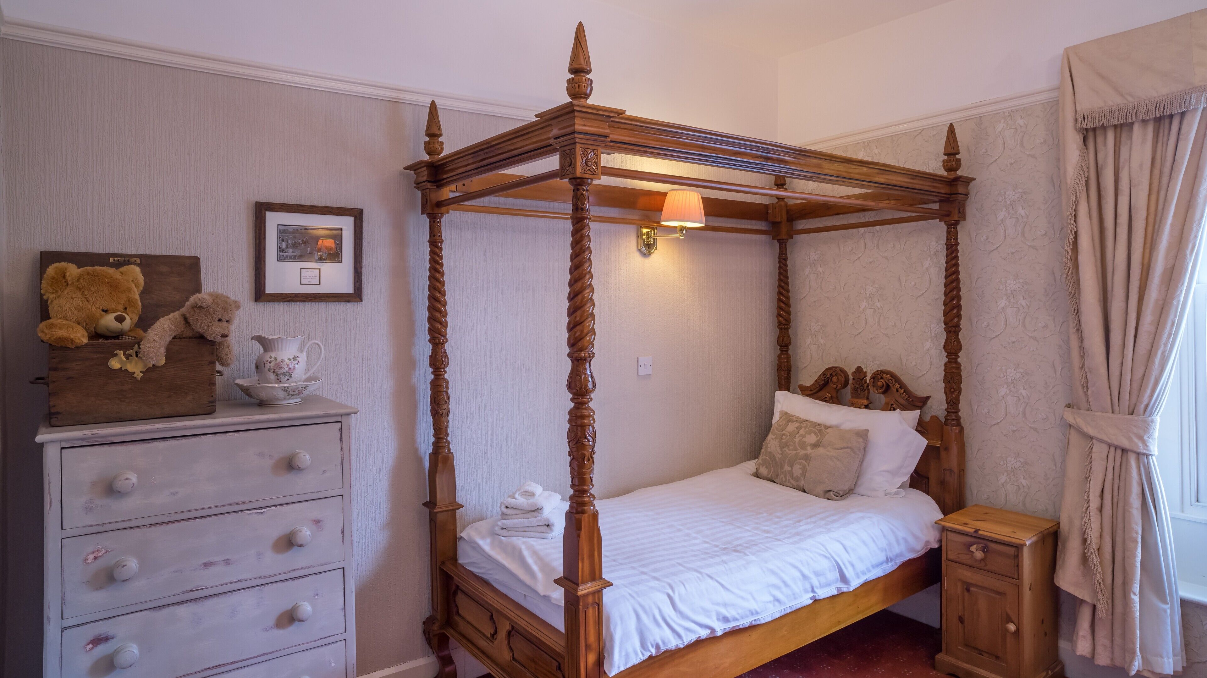 A four-poster canopy bed with a wooden finish, a bedside table, a dresser with multiple drawers, and a framed picture on the wall.