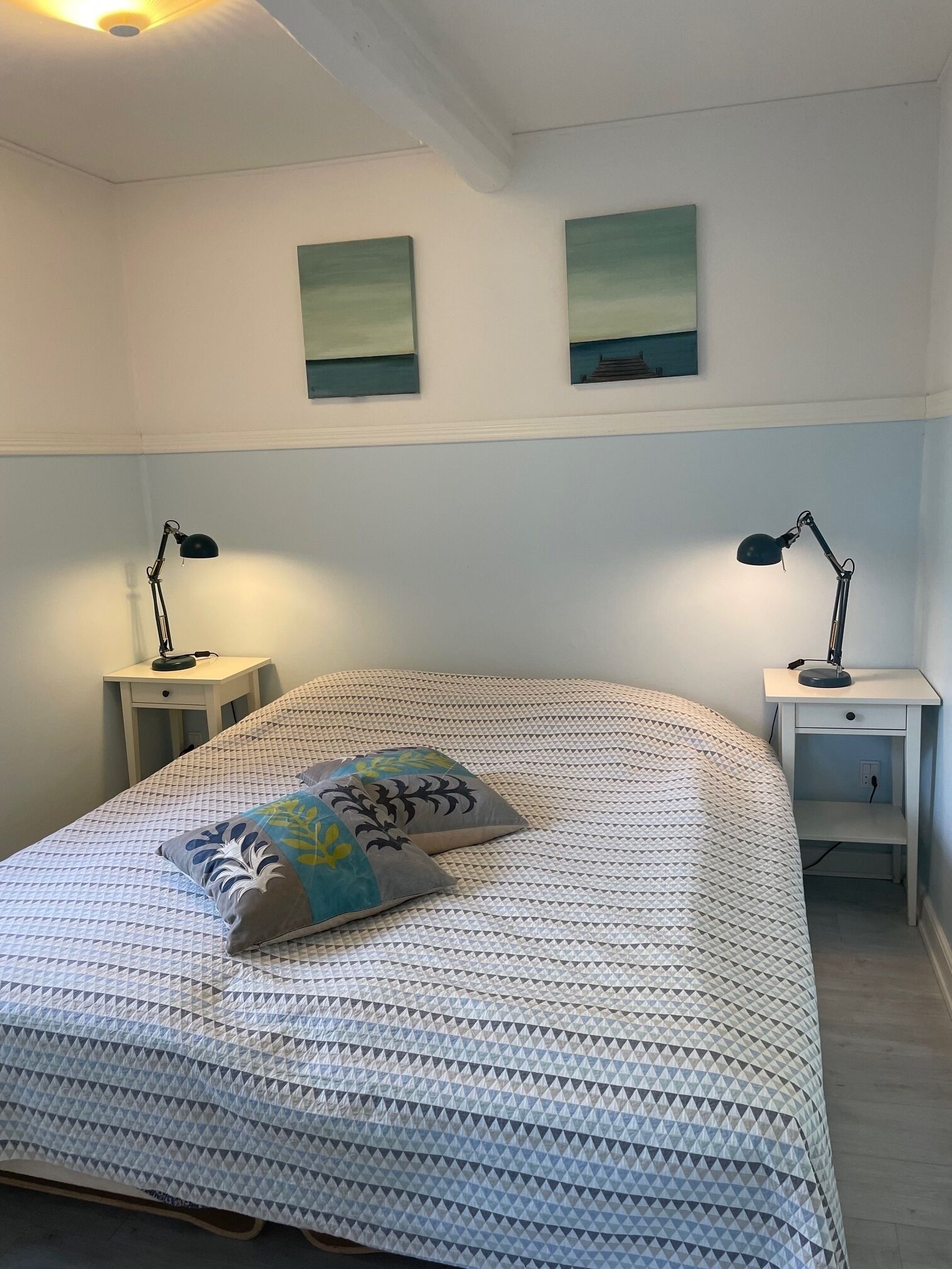 A neatly made bed with a patterned bedspread, two bedside tables with lamps, and two wall-mounted paintings.