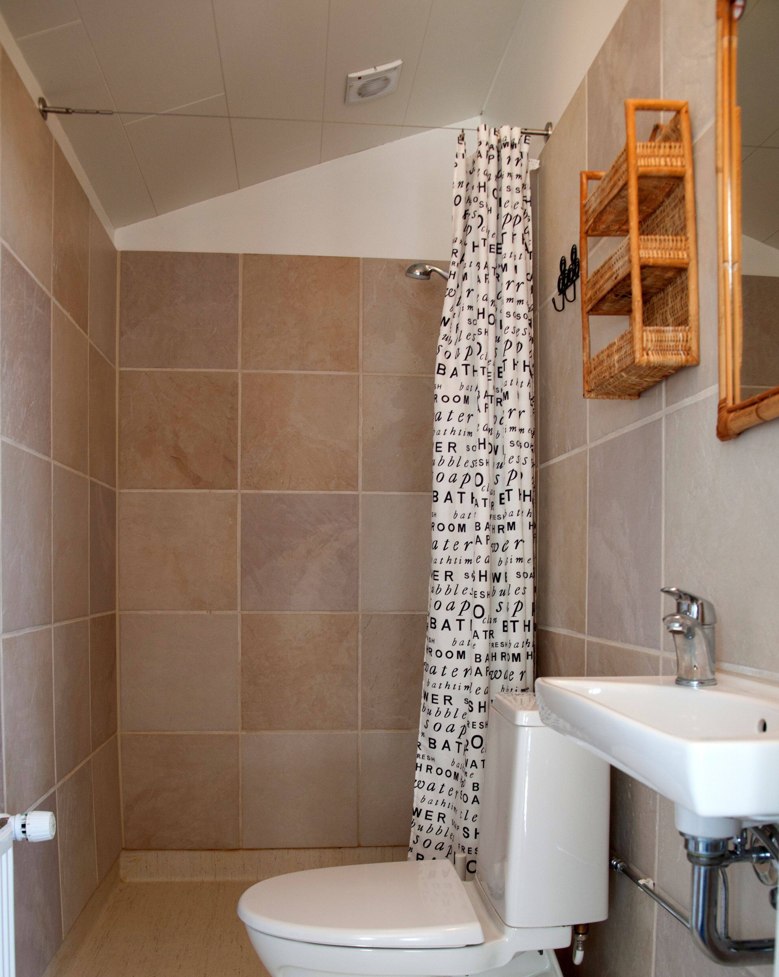 Um banheiro com azulejos bege, vaso sanitário branco, cortina de chuveiro com escrita, prateleira de madeira e um espelho.