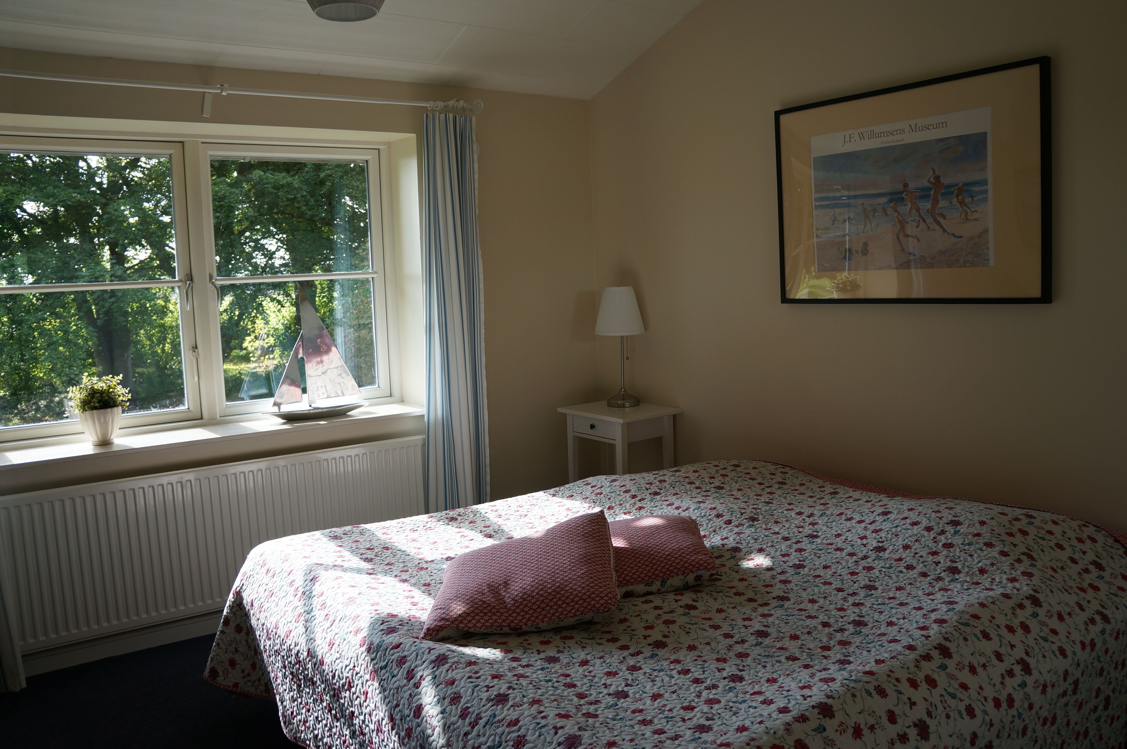 A bedroom with a bed, a window with curtains, a radiator, a small bedside table with a lamp, and a framed picture on the wall.