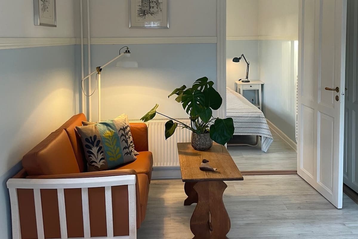 Quarto com um banco branco, uma mesa de madeira, uma planta e uma cama ao fundo.