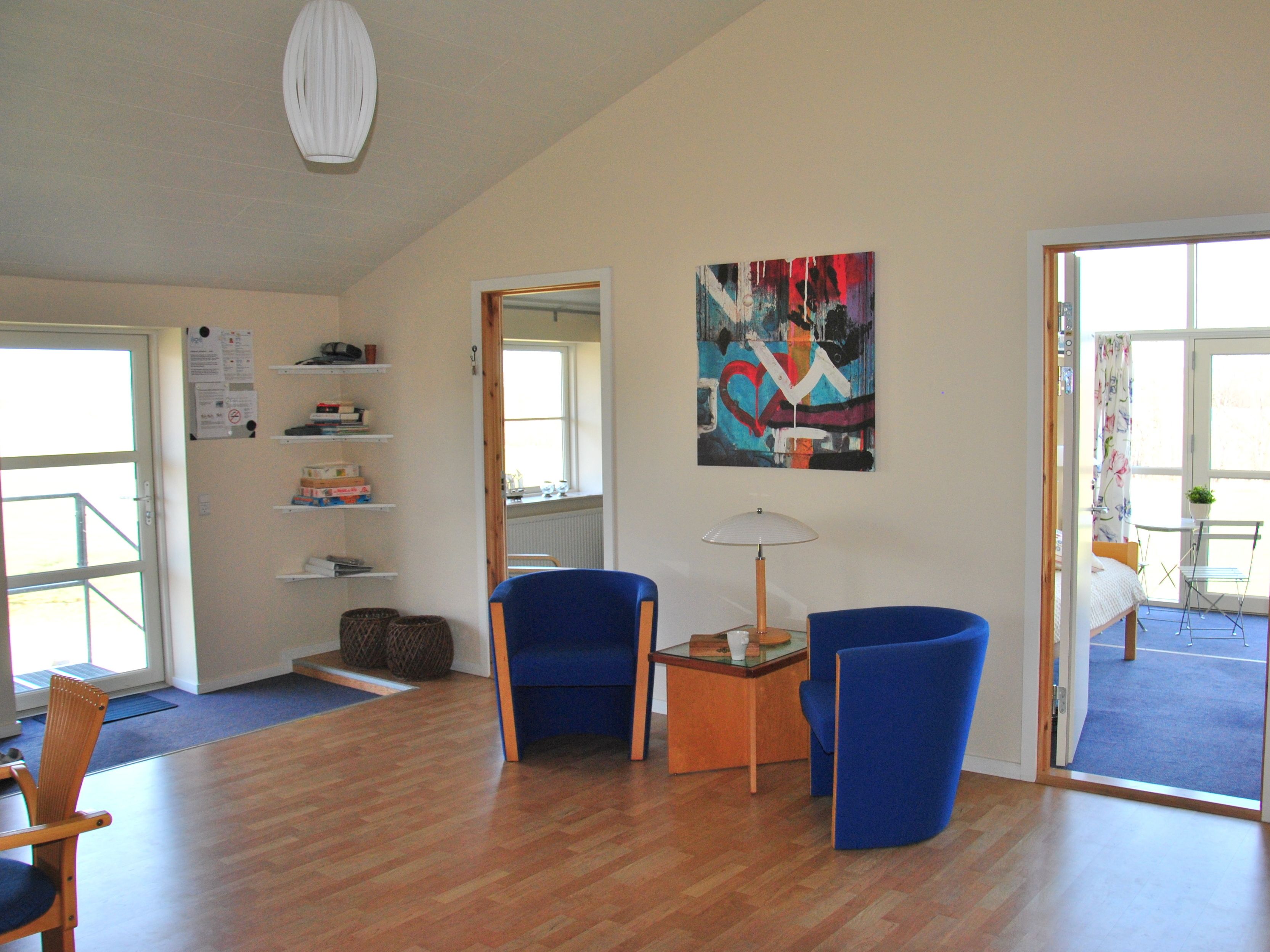 A spacious room with wooden flooring, a blue armchair, a small table with a lamp, and a colorful abstract painting on the wall.