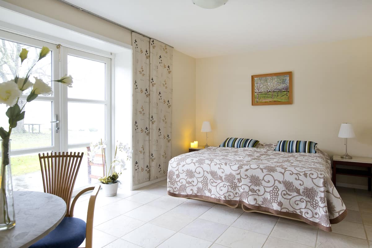 Um quarto com cama, mesa de cabeceira, luminária, cadeira, mesa e janela com cortinas.