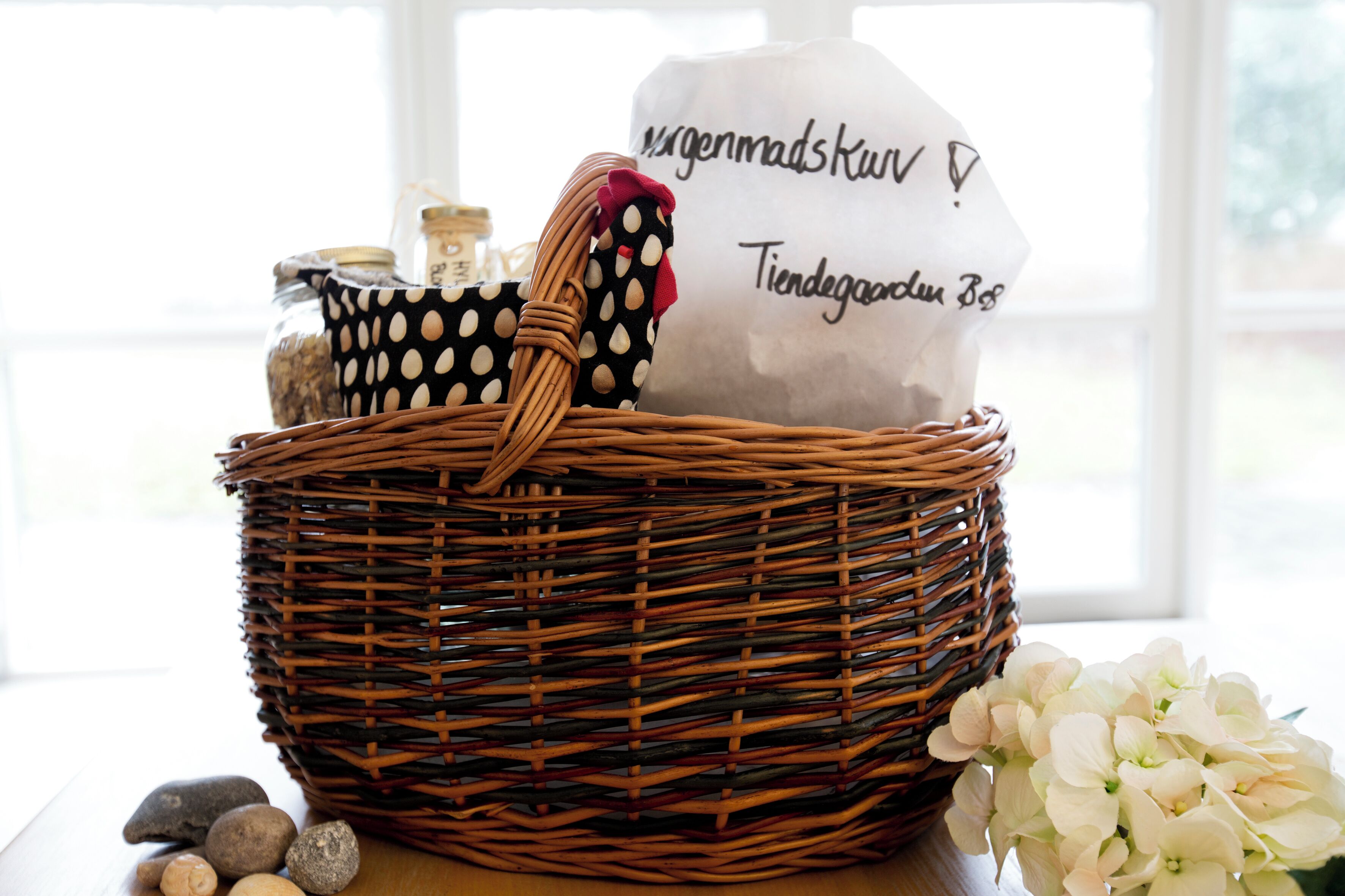 A wicker basket with a polka-dotted cloth and a white bag with text, placed on a wooden surface with rocks and white flowers nearby.