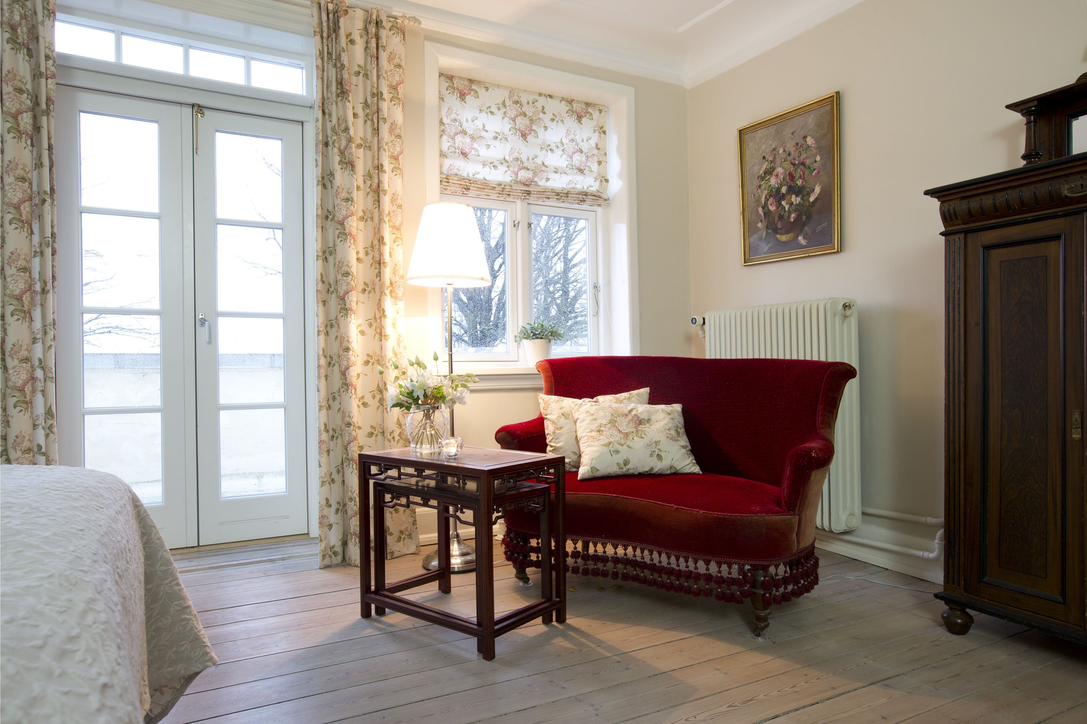 A room with a red velvet sofa, a wooden table, a radiator, and a window with floral curtains.