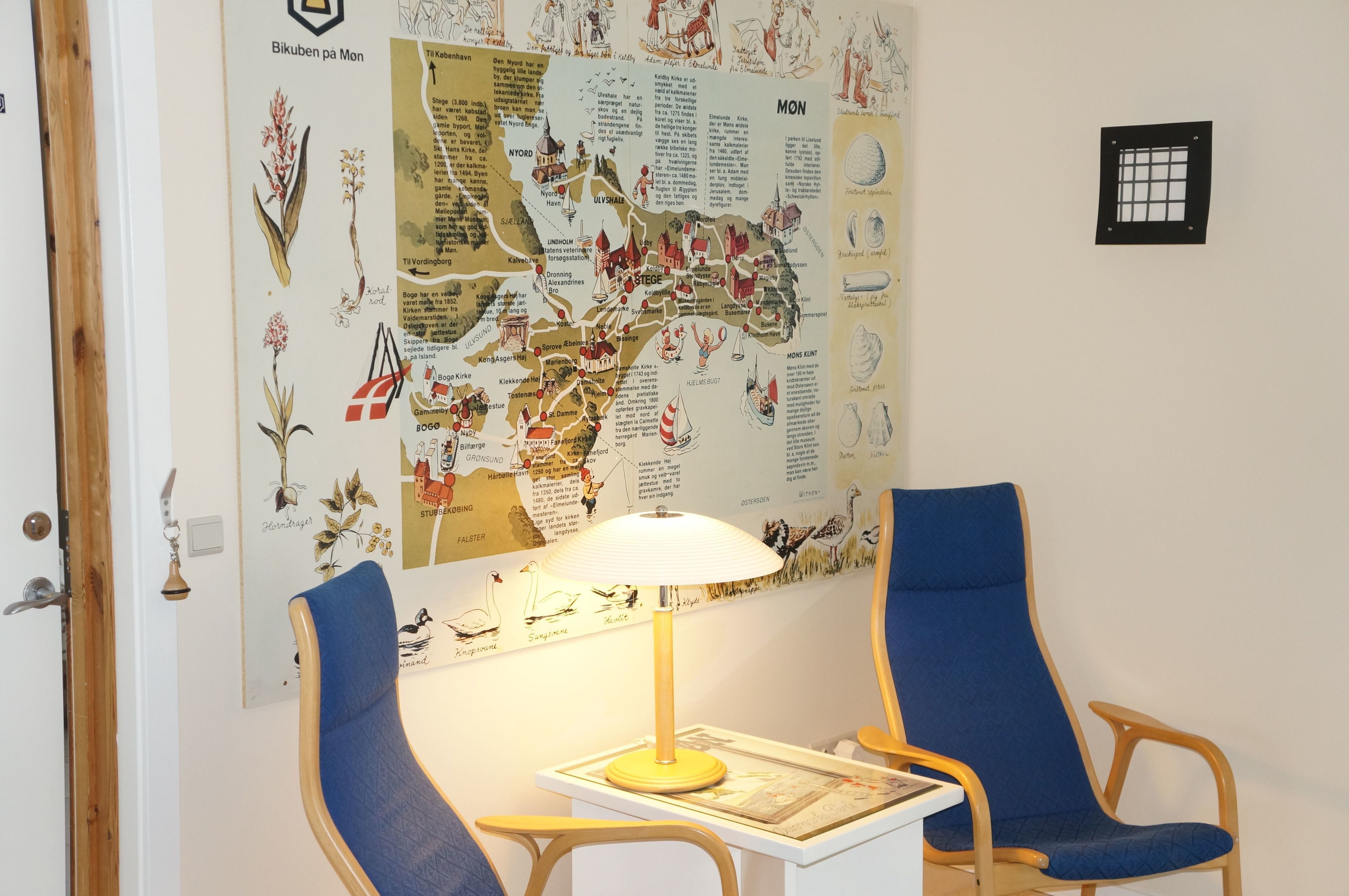 A room with a large map on the wall, two blue chairs, a wooden table, and a lamp.