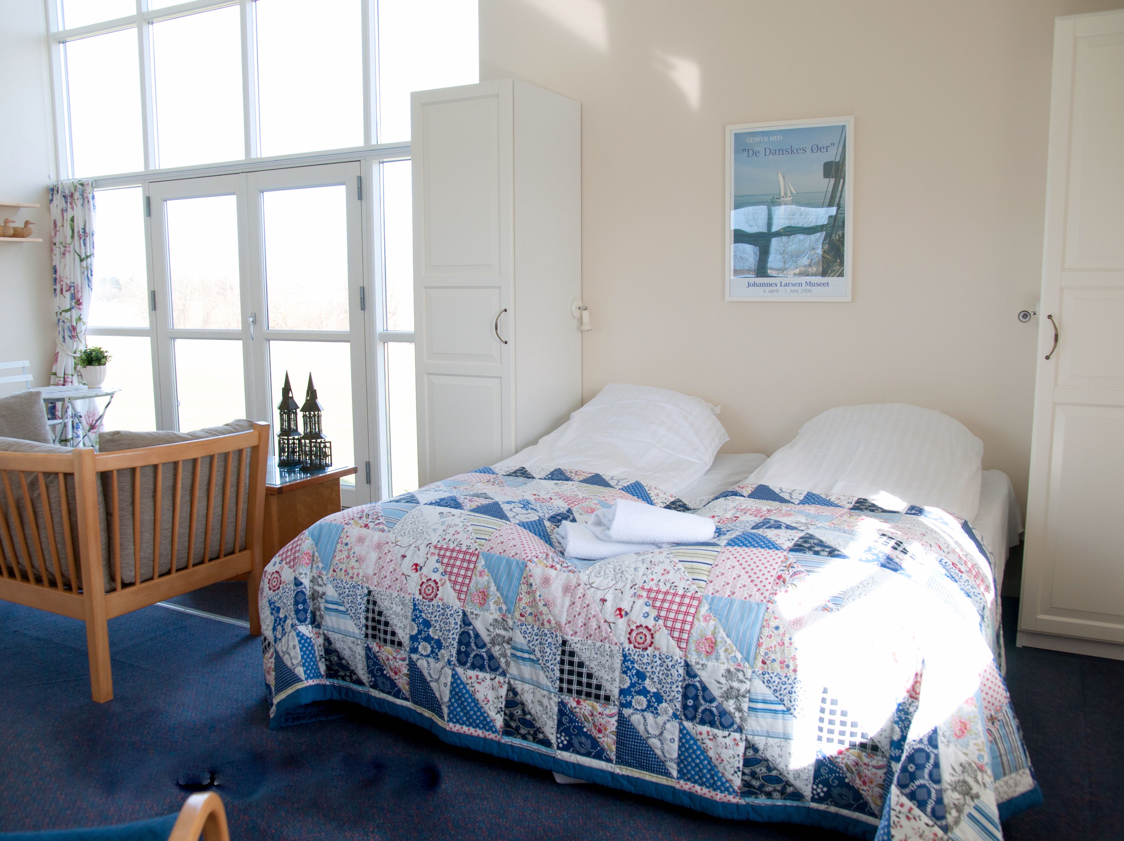 A bedroom with a large bed, a wooden chair, a small table with a plant, and a window with curtains.