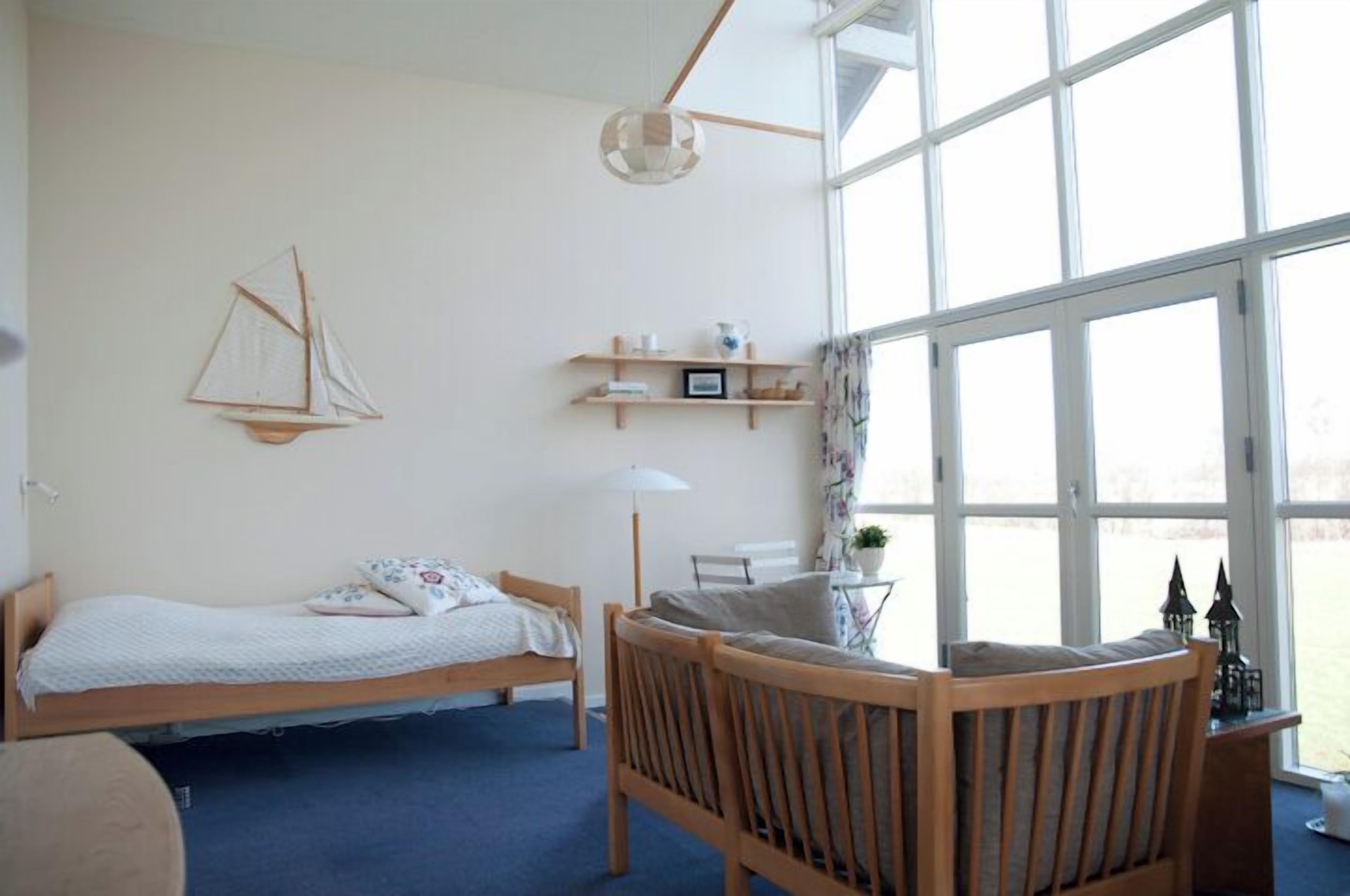 A room with a bed, a wooden chair, a small table, a lamp, and a large window.