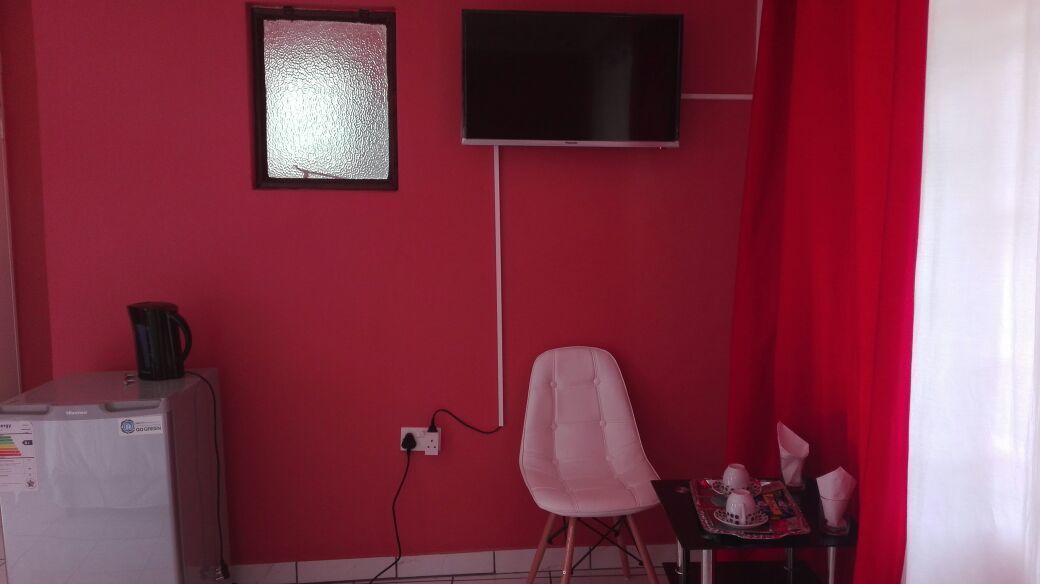 A room with a red wall, a flat-screen TV mounted above a window, a white chair, and a small table with a teapot and cups.
