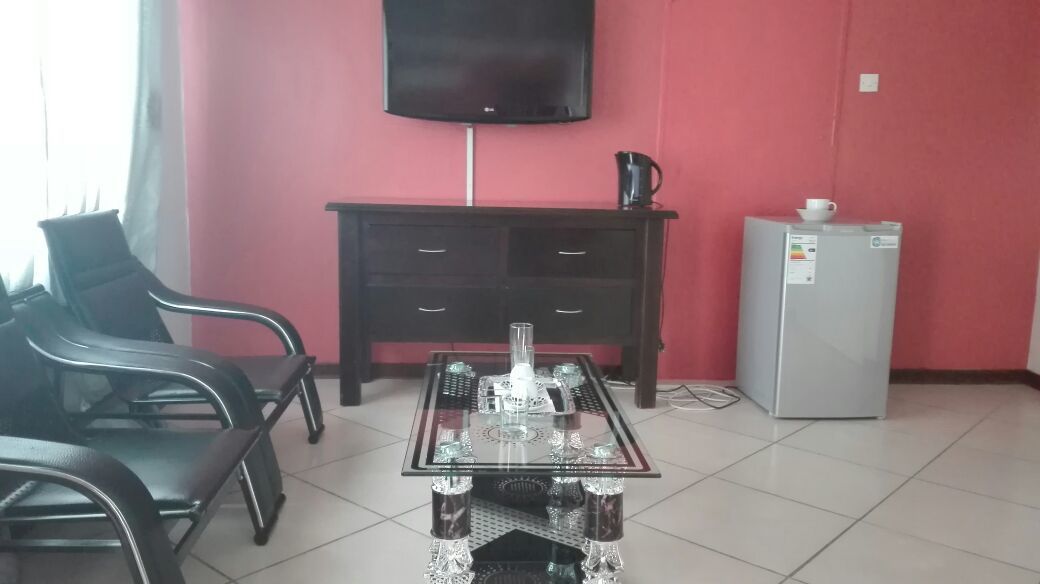 A room with a glass-top table, black chairs, a dark wooden cabinet, a television mounted on the wall, and a refrigerator.
