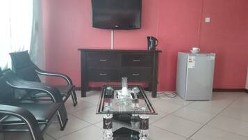 A room with a glass-top table, black chairs, a dark wooden cabinet, a television mounted on the wall, and a refrigerator.