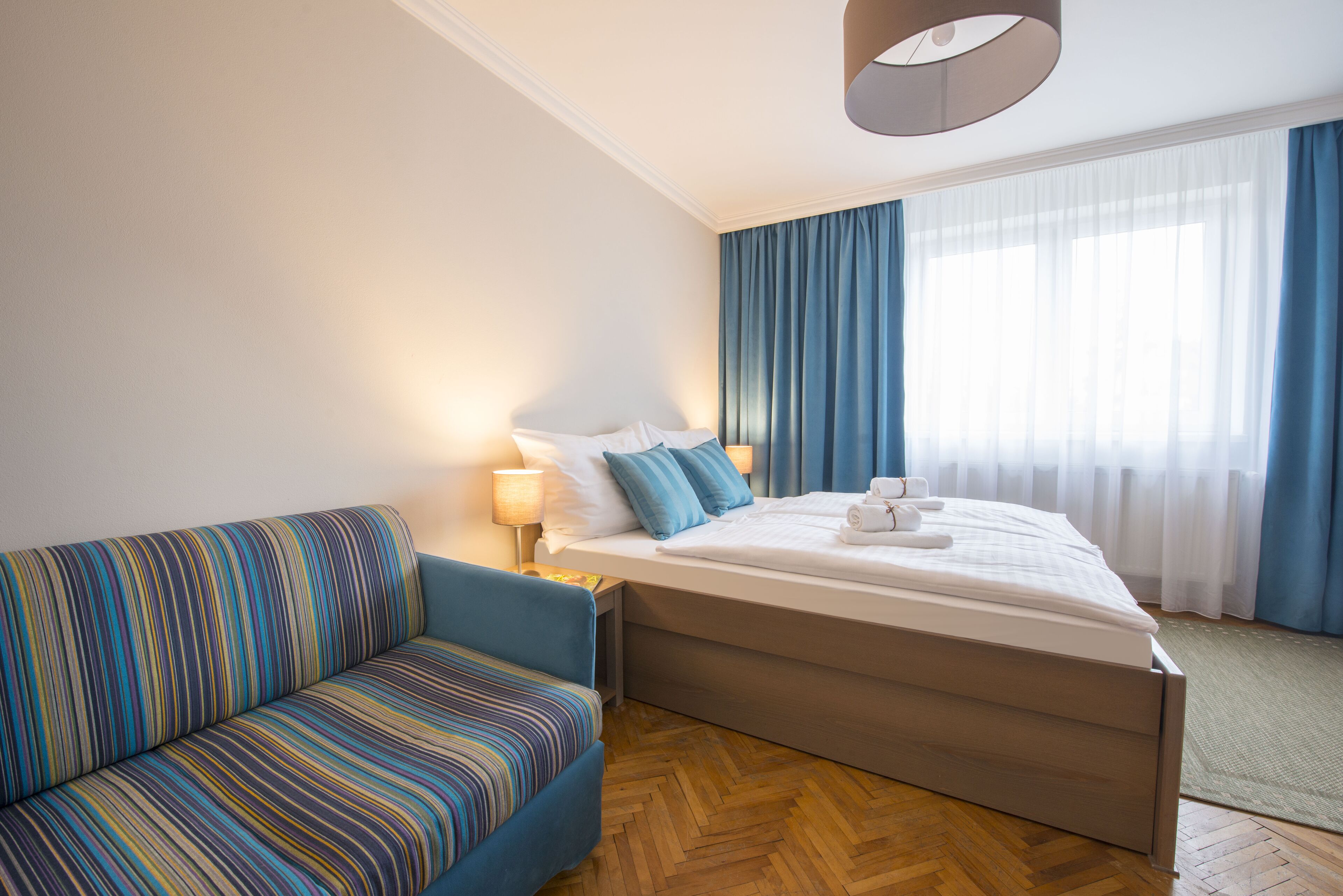 Hotellihuone, jossa on sänky, sohva, lamppu ja ikkuna, jossa on verhot.