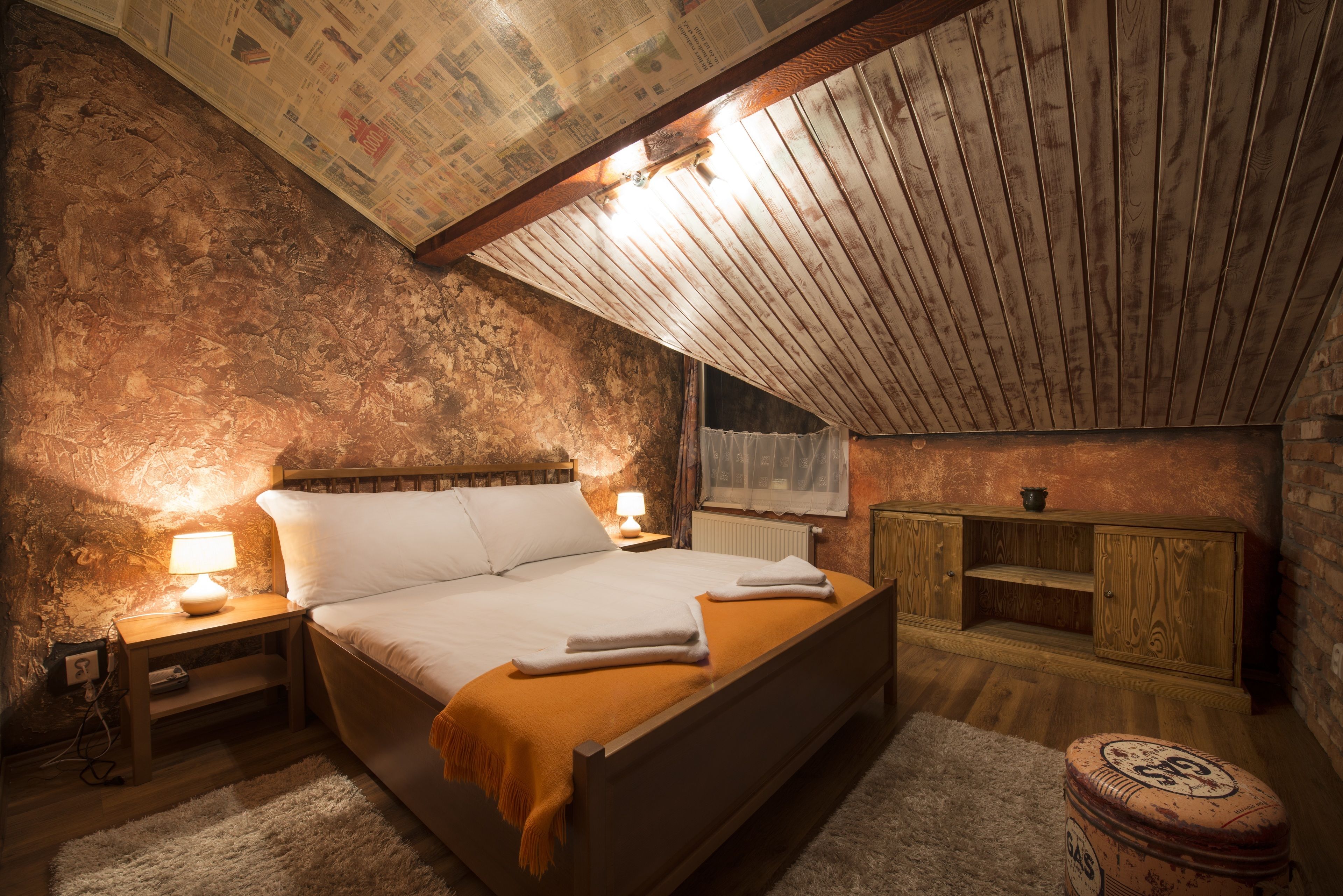 Camera da letto con soffitto spiovente, testiera in legno, un letto con lenzuola bianche, comodino con lampada, armadio in legno e termosifone.