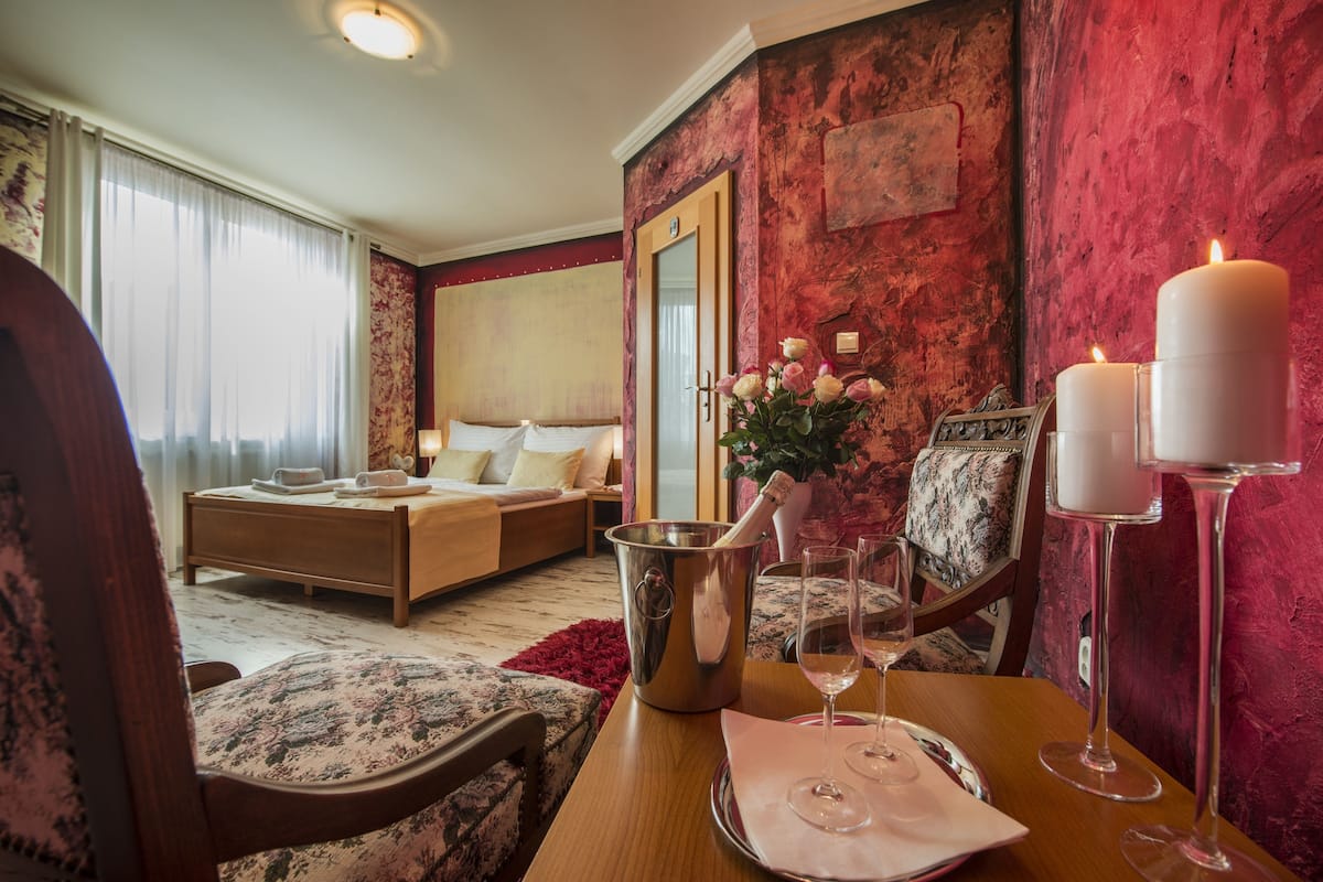 A hotel room with a bed, a sitting area with a table set for two, a vase with flowers, and two lit candles on the table.