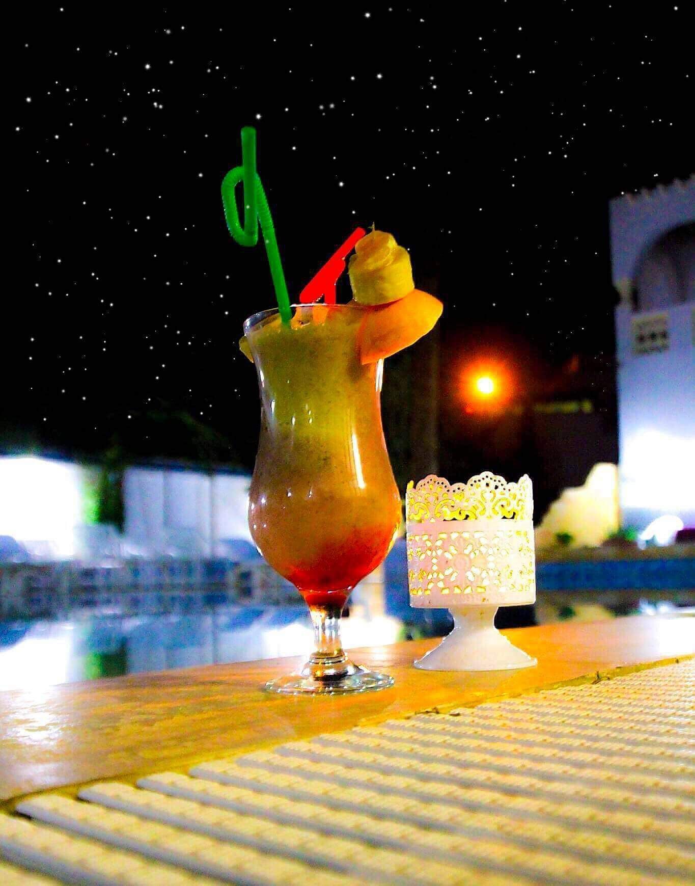 Un cocktail servi dans un grand verre, avec une paille et une tranche d’ananas sur le bord, et un petit gâteau blanc décoré, posé sur une table.