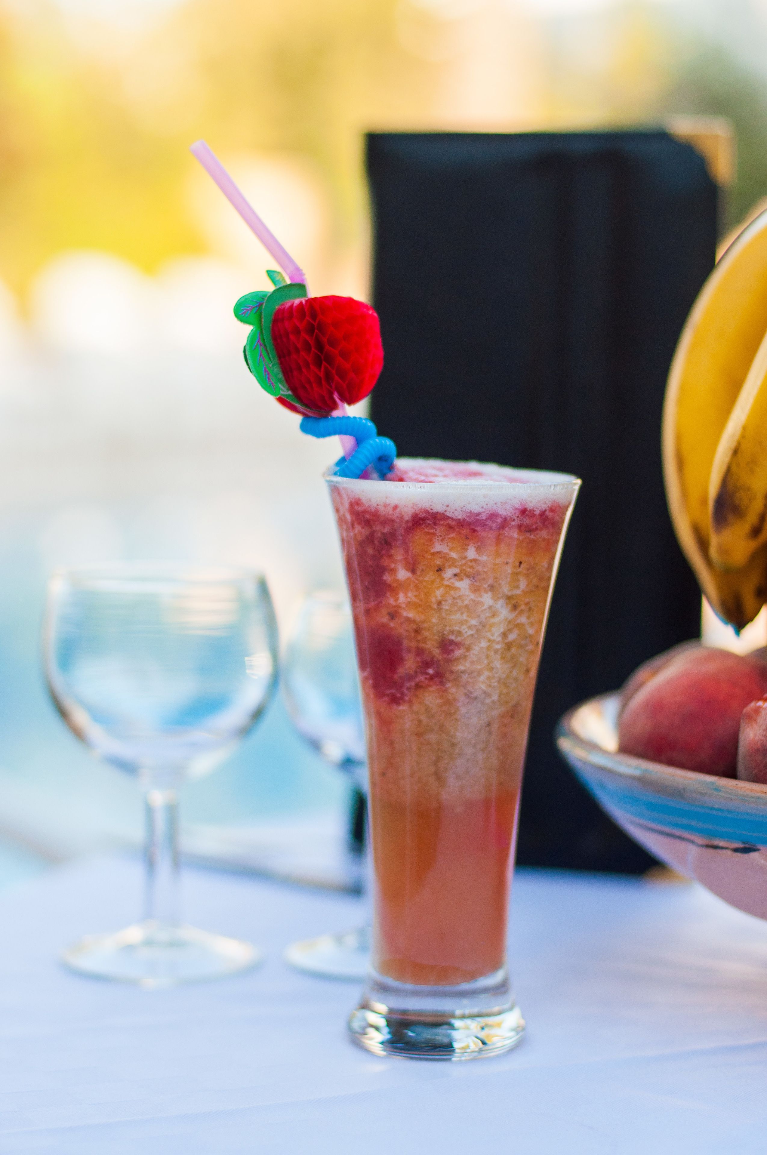 A tall glass with a strawberry and blue straw, placed on a table with a bowl of fruit and a wine glass in the background.