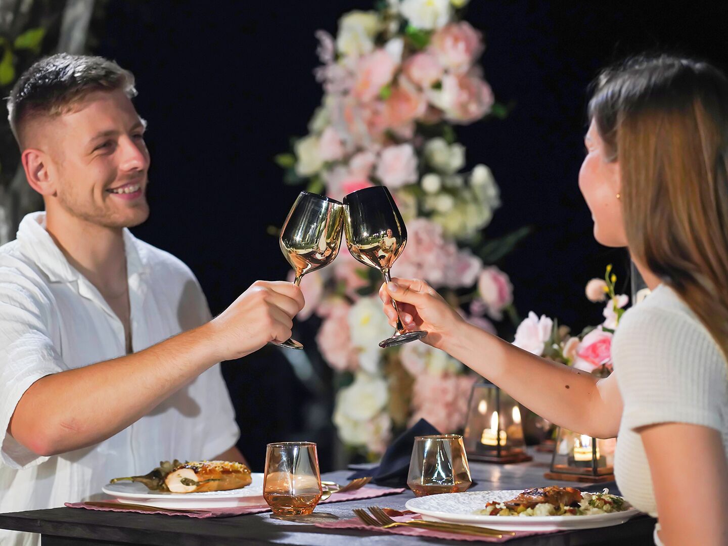 A couple toasting with wine glasses at a table with food and candles.