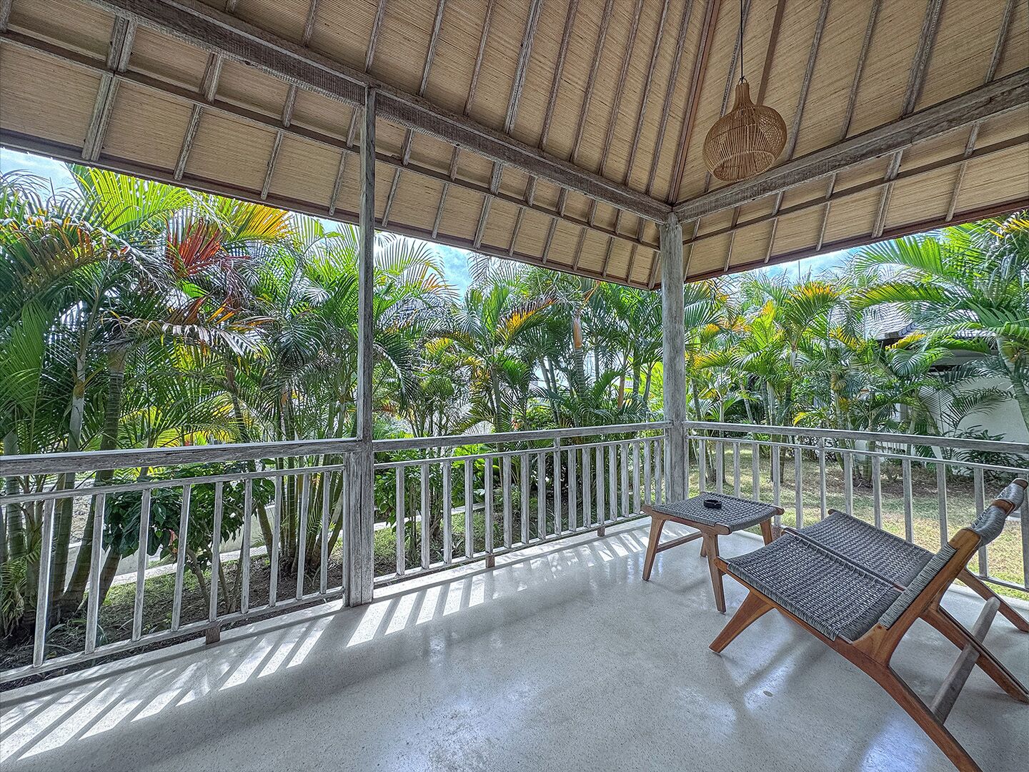 Un espacio al aire libre cubierto con piso de concreto, dos sillas de madera con asientos de mimbre y barandilla de metal, con vegetación tropical exuberante al fondo.