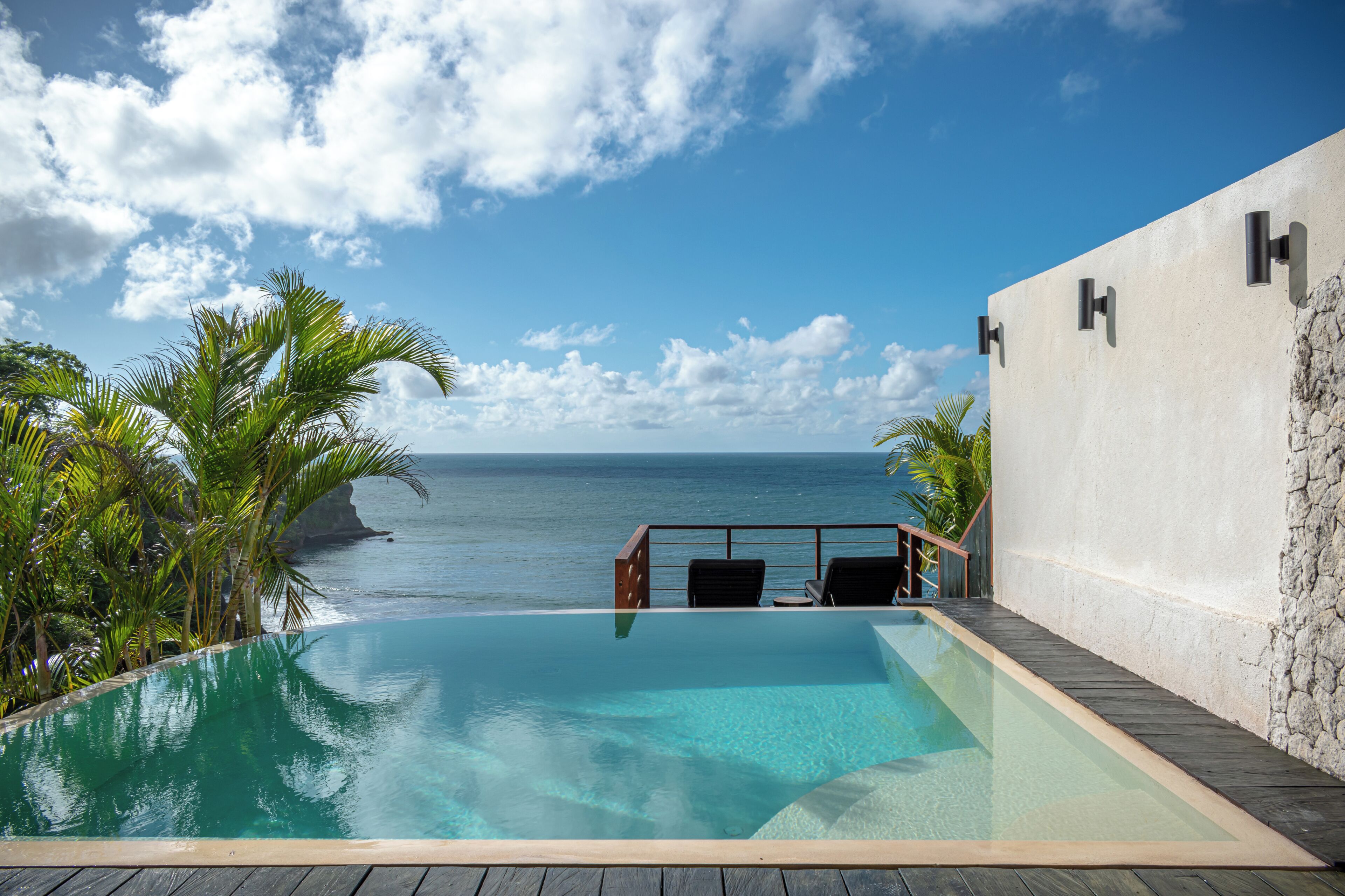 An infinity pool with a view of the ocean, a stone wall, and a wooden deck with chairs.