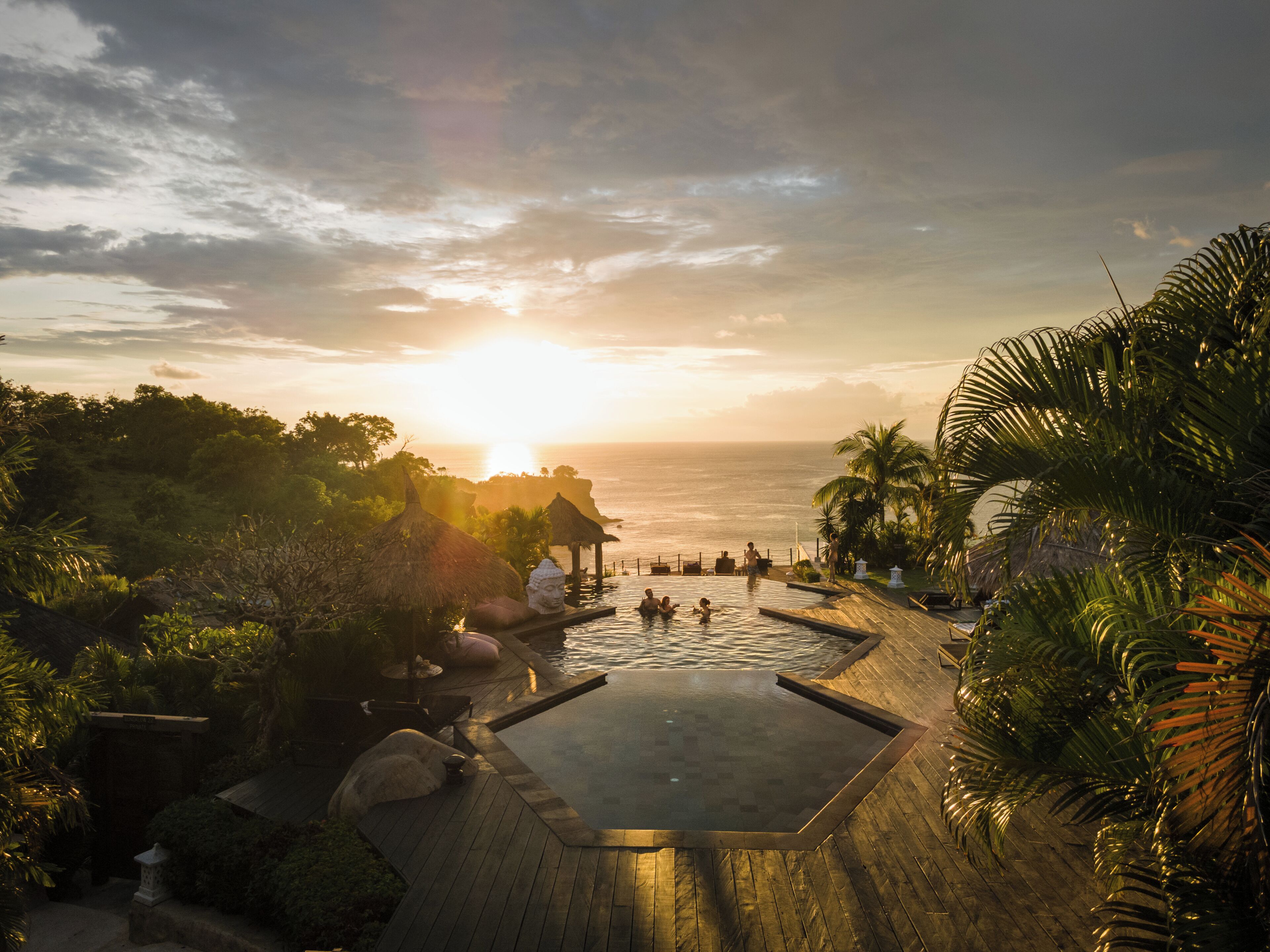 Piscina de resort con vista al atardecer, rodeada de palmeras y una terraza de madera.