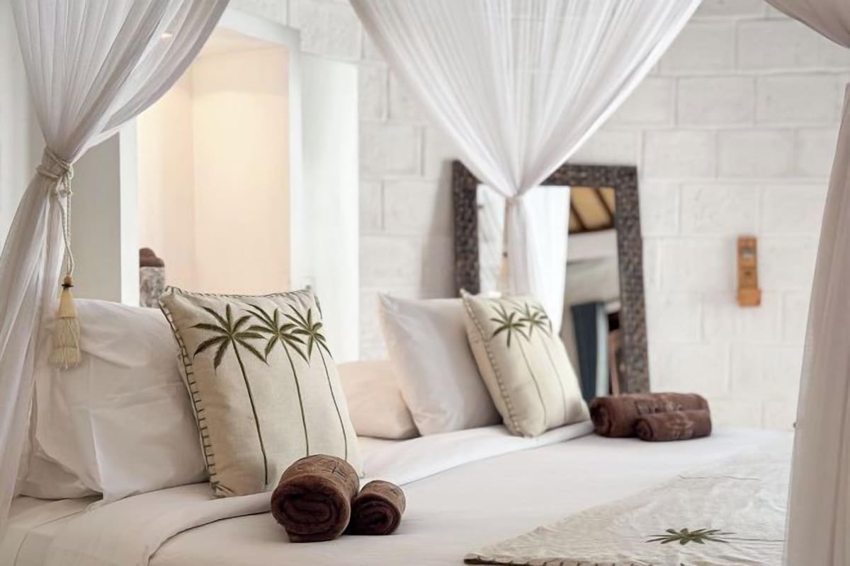 A bed with white linens, a canopy, and palm leaf pillows.