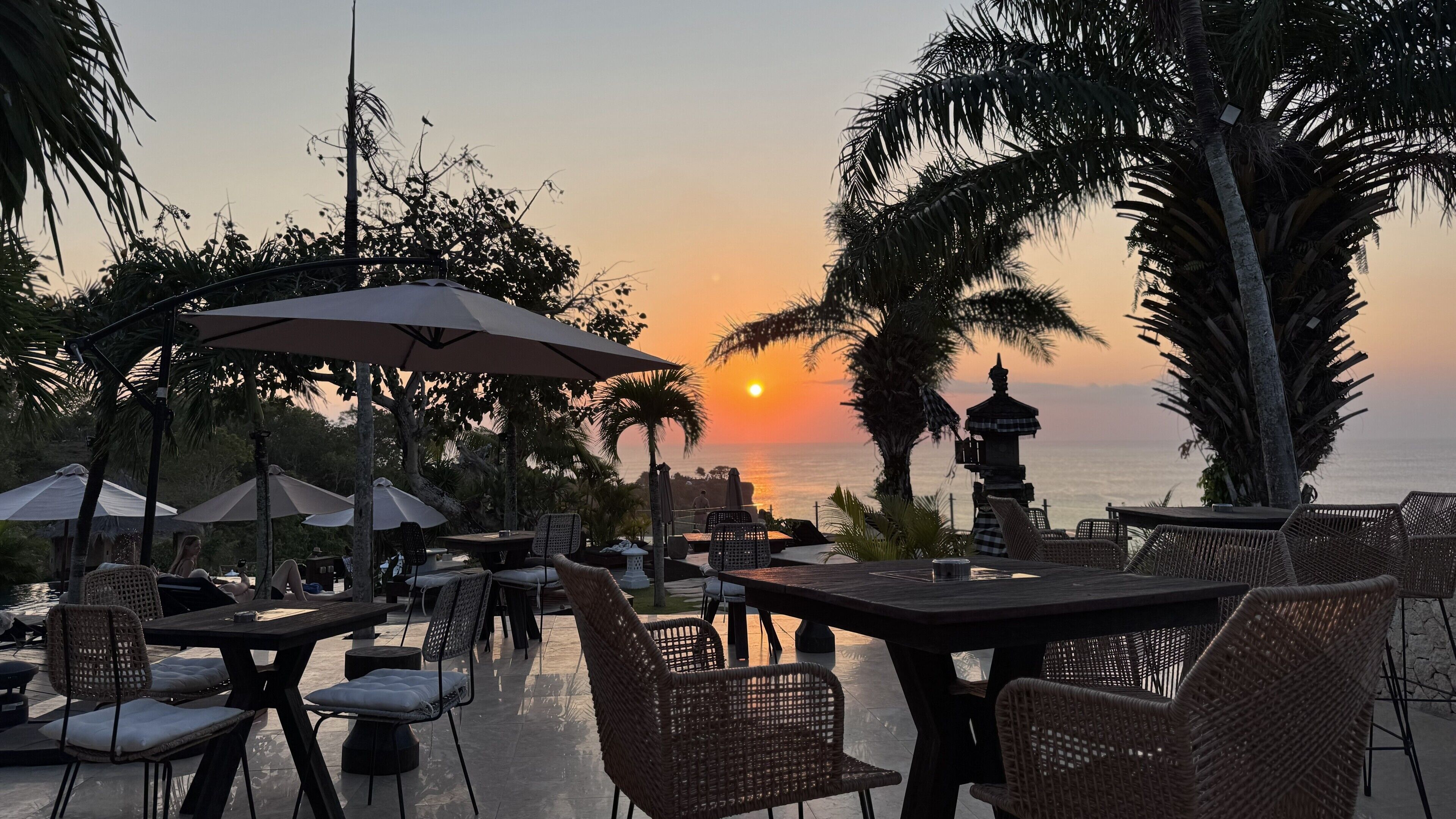 An outdoor seating area with tables and chairs, umbrellas, and a view of the sunset over the water.