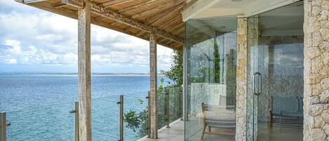 A modern outdoor balcony with a stone wall, glass door, and a view of the sea.