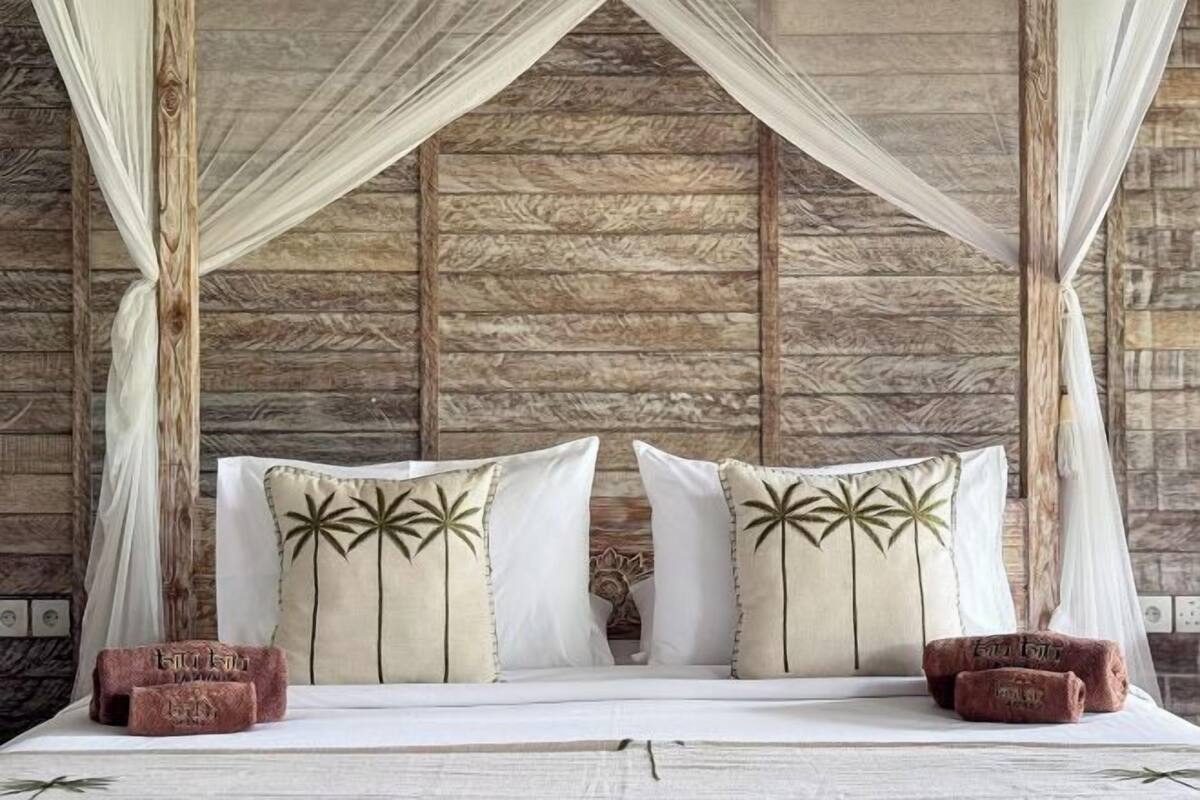 Een bed met wit beddengoed en twee decoratieve kussens met palmboommotieven, voor een rustieke houten wand.