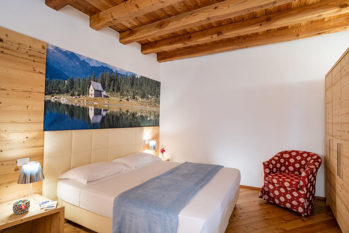 A bedroom with a bed, a wooden headboard, a painting of a lake, a red patterned chair, and a wooden floor.