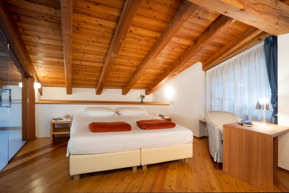 A bedroom with a wooden ceiling, a bed with white bedding, a nightstand, a bedside table with a lamp, and a window with curtains.