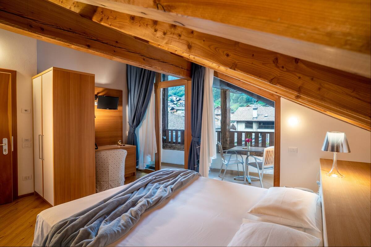 A bedroom with a wooden ceiling, a bed with white linens, a wooden wardrobe, and a balcony with a table and chairs.