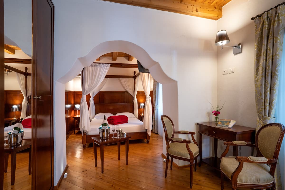 A bedroom with a four-poster bed, a bedside table with a lamp, a chair, and a small desk with a lamp.