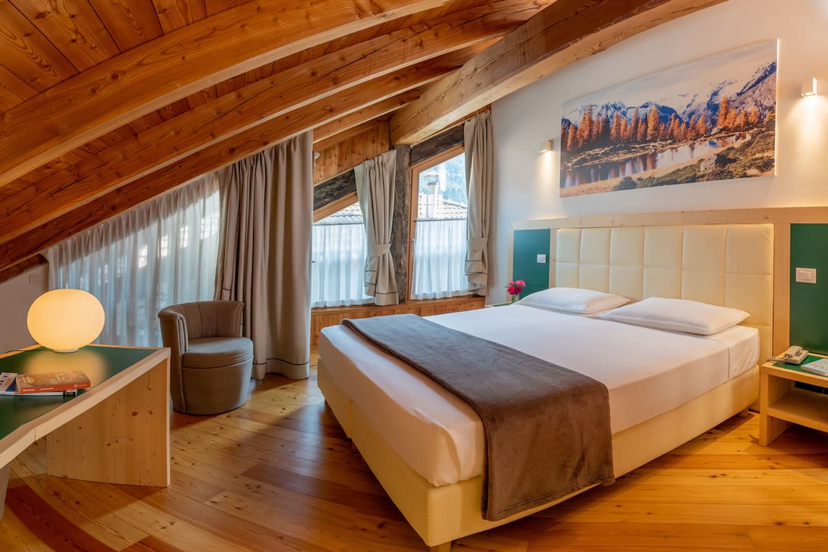 A cozy room with a wooden ceiling, a bed with a white headboard, a desk with a lamp, and a painting of a mountain landscape.