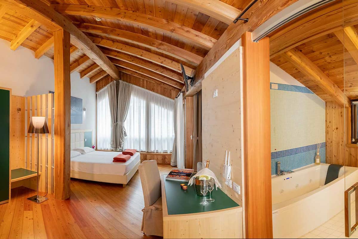 A wooden-floored bedroom with a bed, a desk with a drink, and a bathtub.