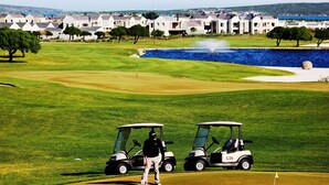 A golf course with two golf carts, a pond, and a row of houses in the background. - Golf Paradise, Langebaan 6-sleeper (Langebaan)