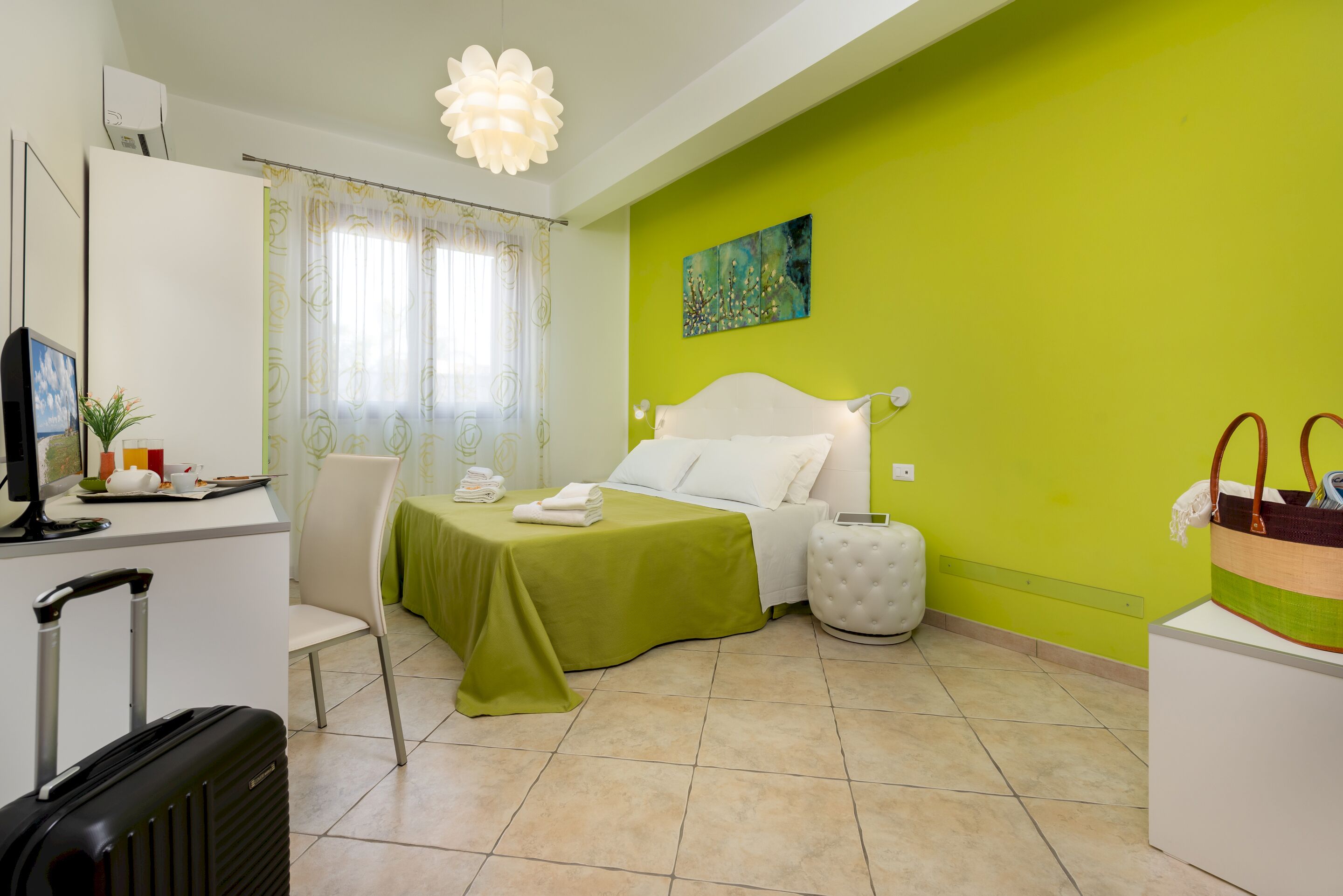 A hotel room with a bed, a desk, a chair, a suitcase, and a painting on the wall.