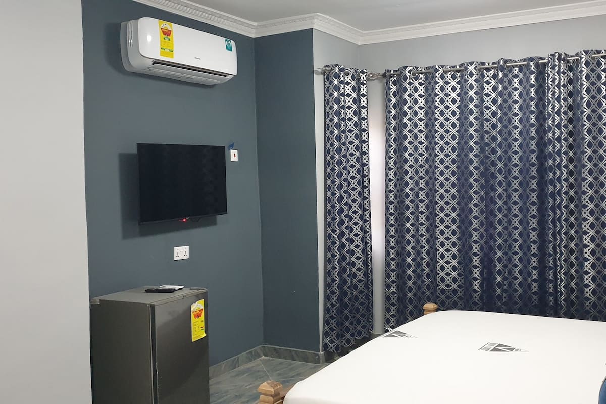 Ein Schlafzimmer mit einem Bett, einem an der Wand montierten Fernseher, einer Klimaanlage und einem gemusterten Vorhang.
