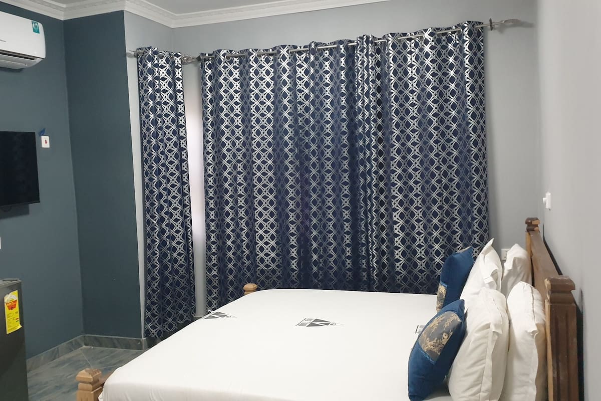 Ein Schlafzimmer mit Bett, einem flachbildfernseher an der Wand, einem Kühlschrank und einem gemusterten Vorhang.