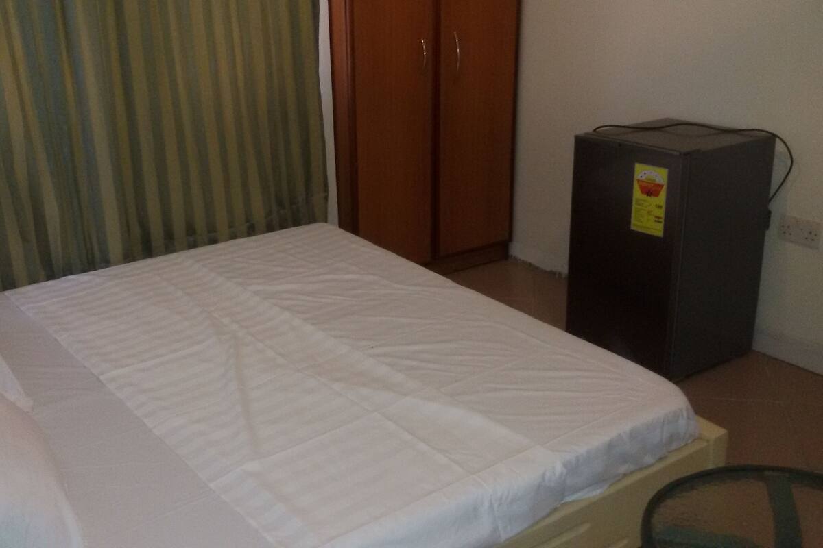 Ein Einzelbett mit weißer Matratze und Kissen, ein Holzschrank und eine kleine, schwarze Kommode mit Aufkleber.