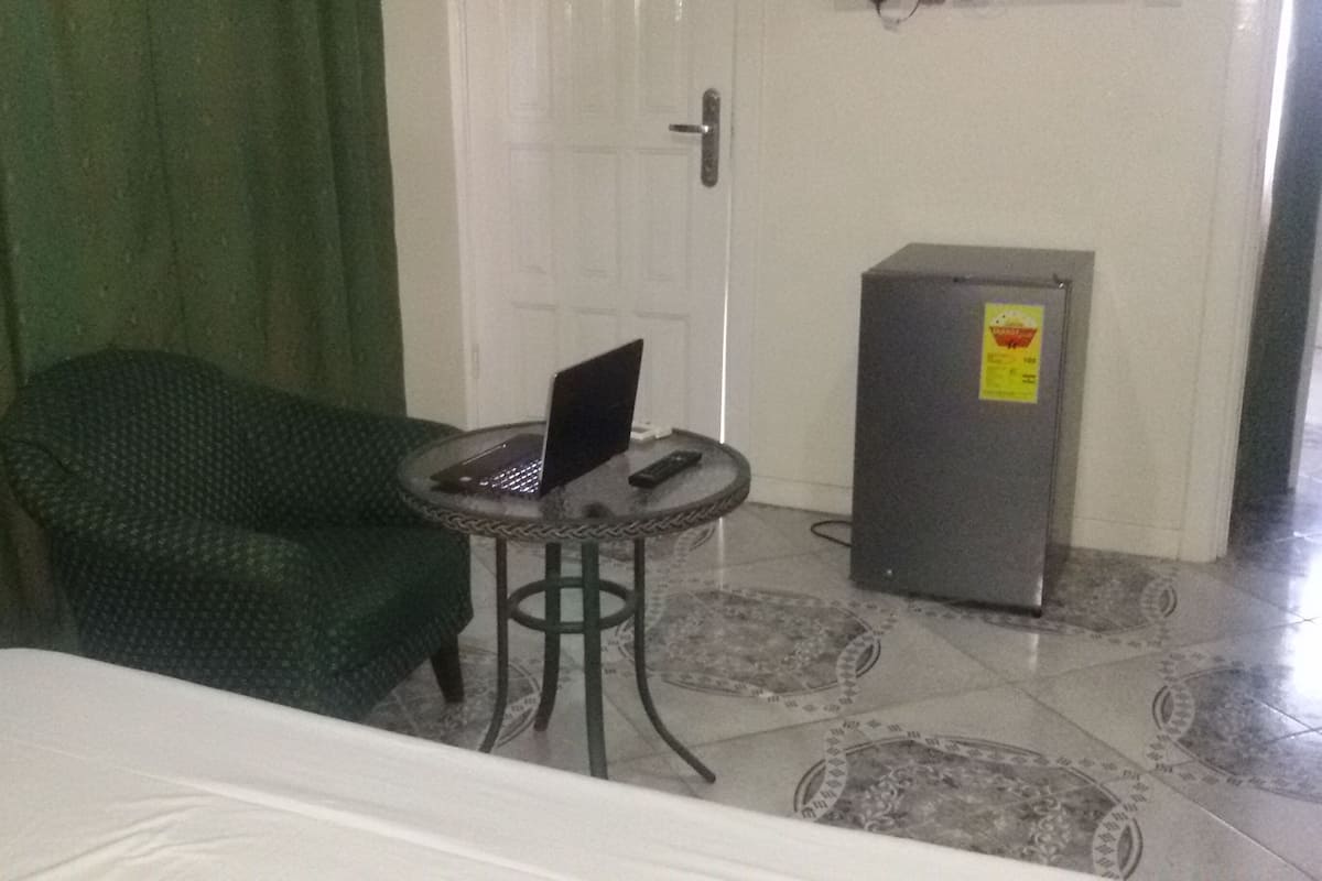 Ein Zimmer mit einem Bett, einem grün gemusterten Sessel, einem kleinen runden Tisch mit einem Laptop und einem Kühlschrank.