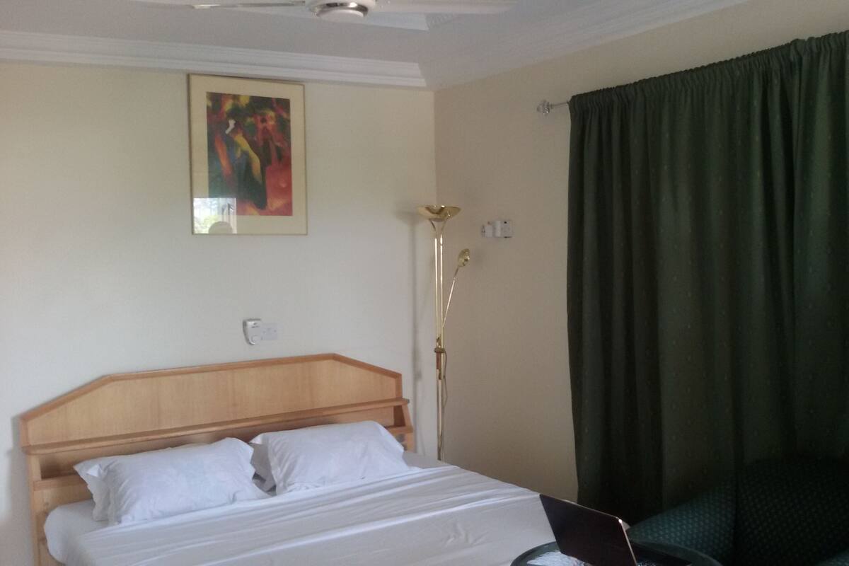 Ein Hotelzimmer mit Bett, Schreibtisch, Lampe, einem Bild an der Wand und einem Fenster mit Vorhängen.