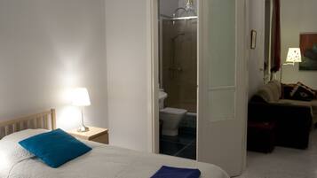 Une chambre avec un lit, une table de chevet avec une lampe, une salle de bain visible à travers une porte ouverte, et un canapé dans le salon attenant.