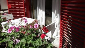 Une table blanche, ornée de fleurs roses, se trouve devant une fenêtre à volets rouges.