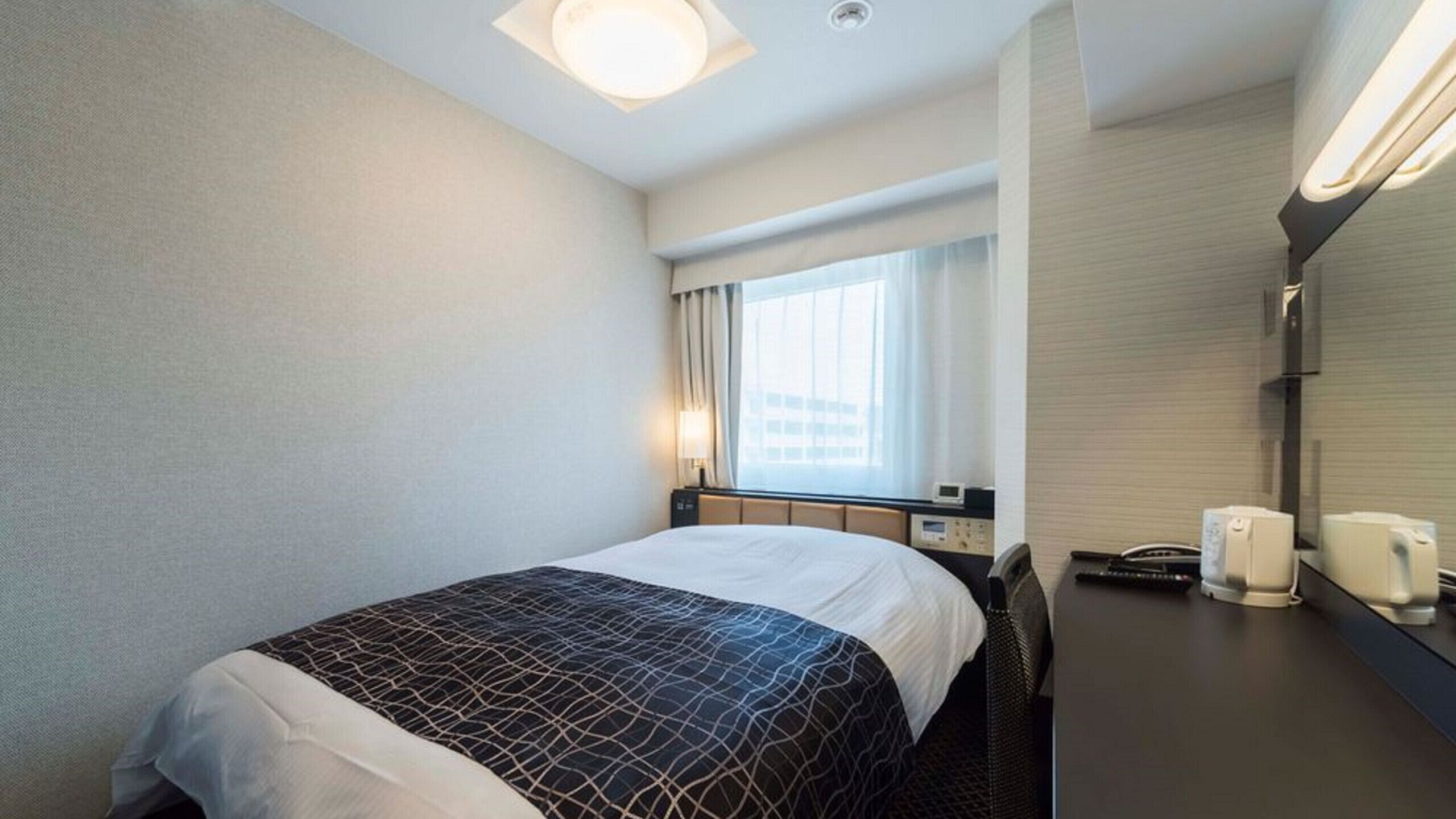Quarto de hotel com cama, mesa de cabeceira, luminária, janela com cortinas e telefone fixado na parede.