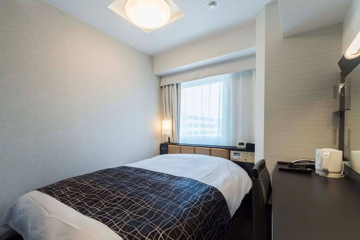 Hotellihuone, jossa on sänky, yöpöytä, lamppu, ikkuna verhoilla ja seinälle asennettu puhelin.
