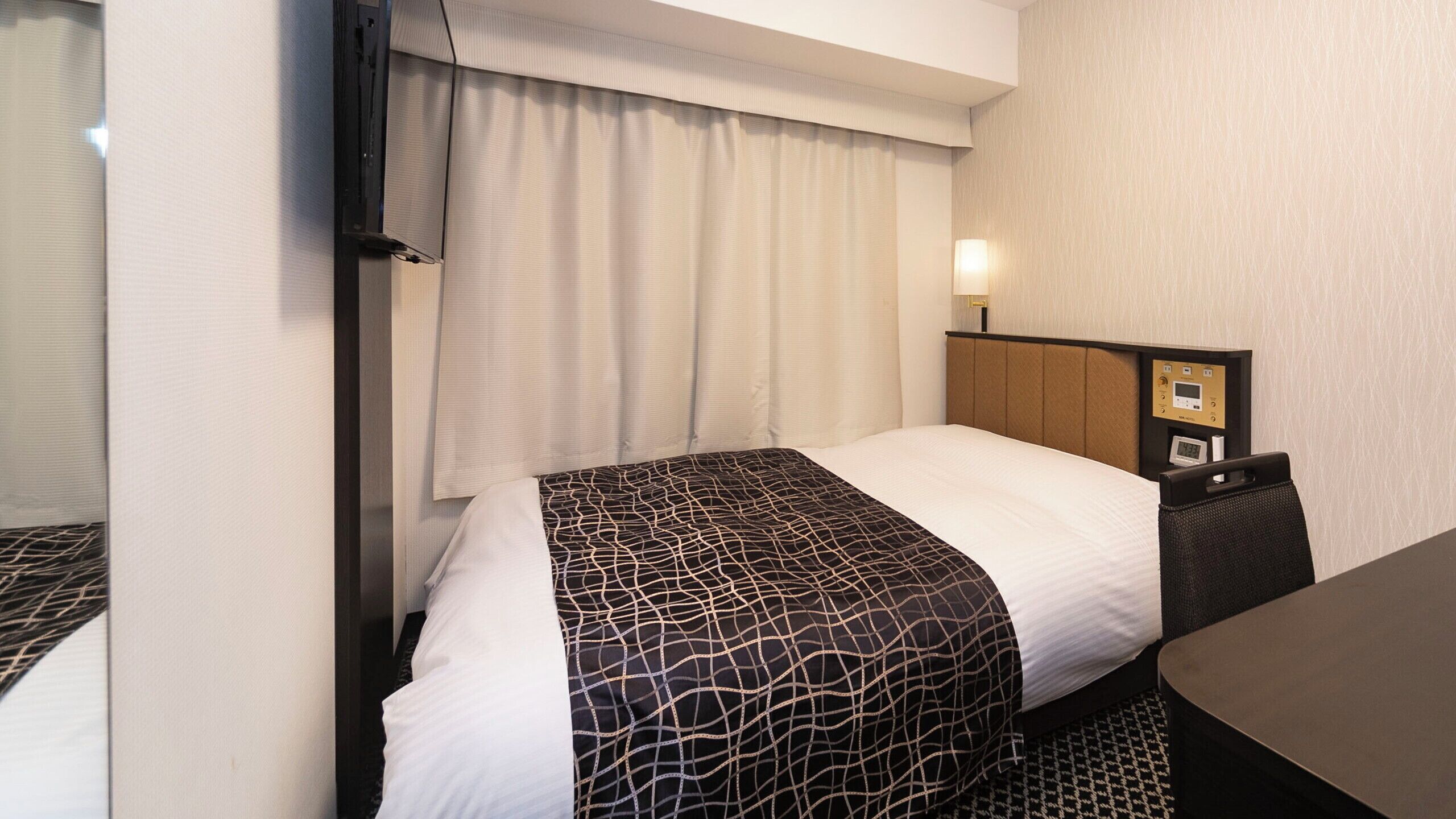 Quarto de hotel com cama, mesa de cabeceira, abajur e painel de controle fixado na parede.