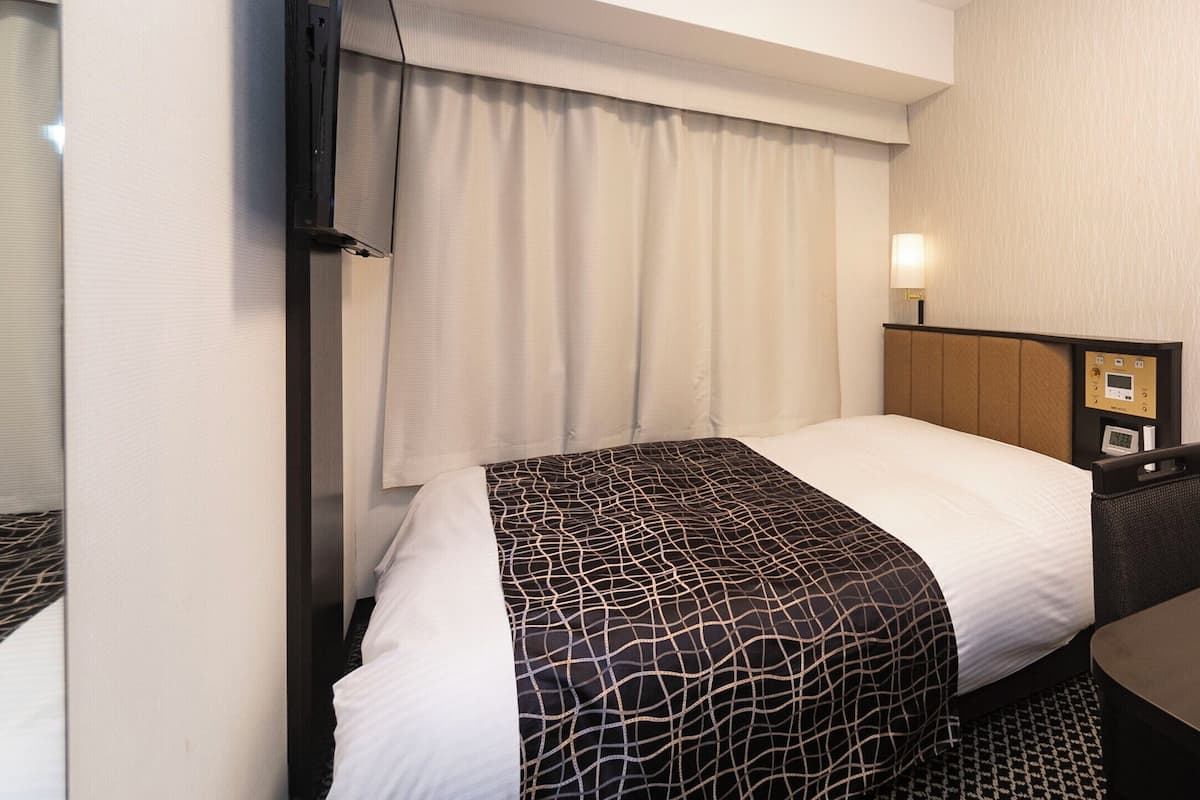 Hotellihuone, jossa on sänky, yöpöytä, lamppu ja seinälle asennettu ohjauspaneeli.