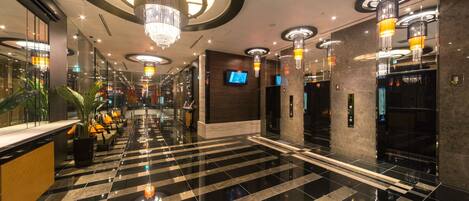 A modern hotel lobby with a checkered black and white floor, multiple chandeliers, and a television on the wall.