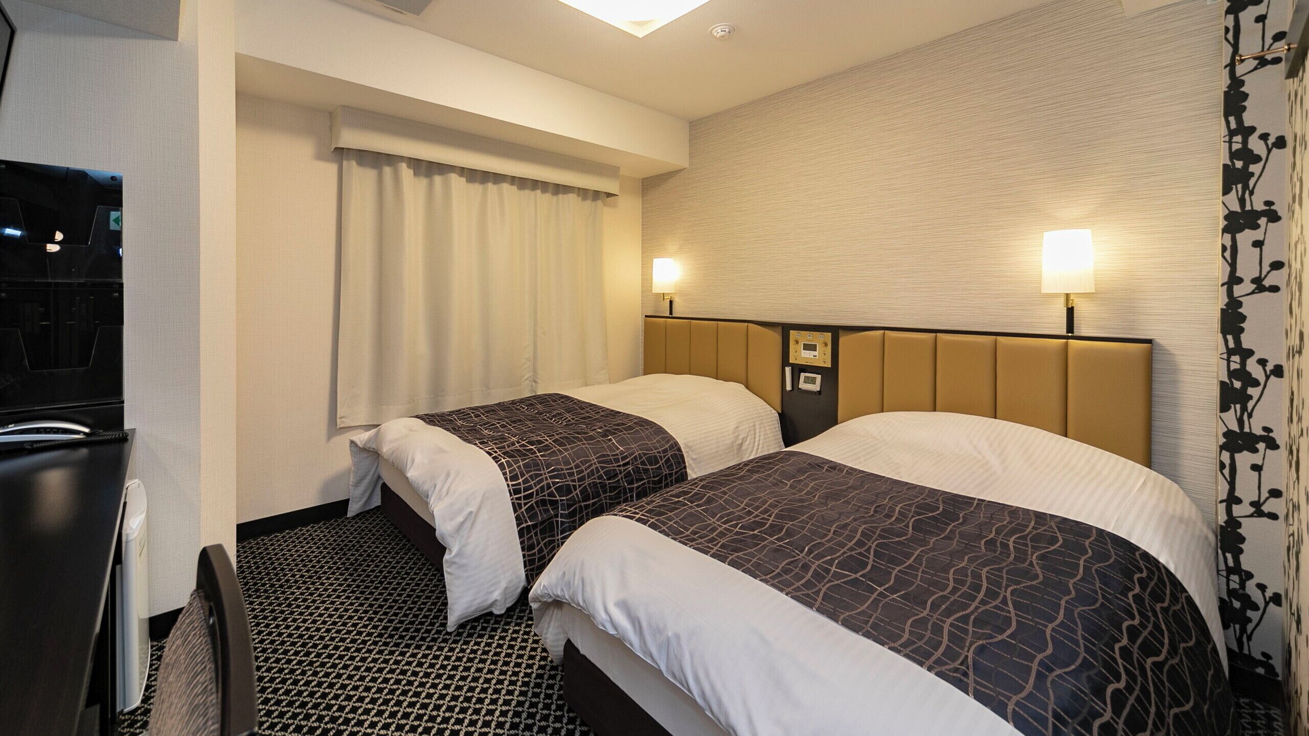 Quarto de hotel com duas camas, cabeceira com painel de controle, janela com cortinas e carpete estampado.
